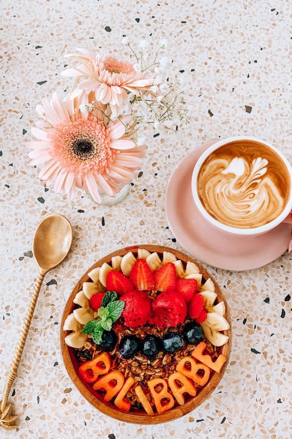 A wooden bowl filled with colorful freshly cut fruit and granola on a terrazzo table top with a pink flower, gold spoon, and freshly made cup of coffee in a pink mug.