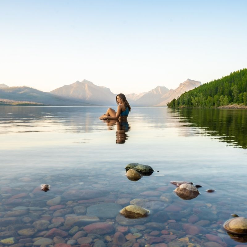 Woman wearing a sports bra sitting in a lake with her upper body out of the water with a beautiful misty mountain range in the distance.