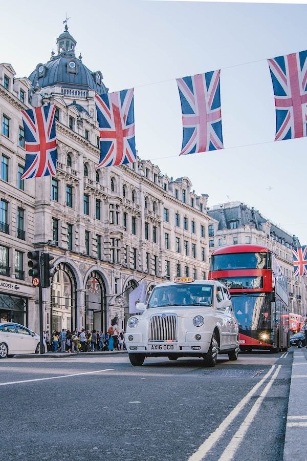 White London taxi in front of a red double-decker bus driving around Piccadilly Circus with British flags in the foreground.