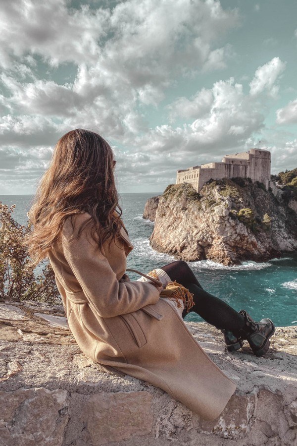 A woman sitting on the walls of Dubrovnik looking out at the Lovrijenac Fortress and Adriatic Sea.