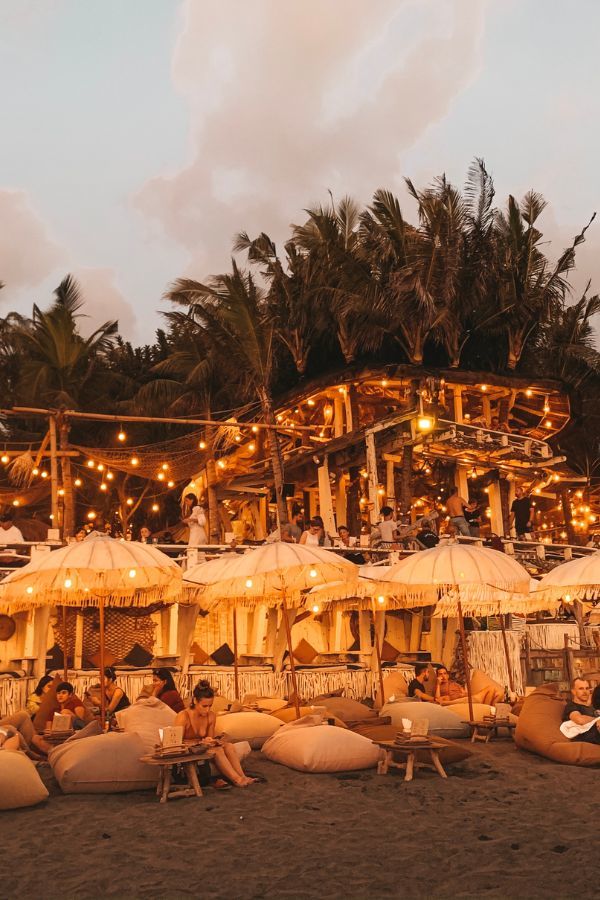 Twilight at a beach club in Bali with cozy bean bags and parasols on the sand, a bustling multi-level wooden bar, all under the glow of string lights and a backdrop of tall palm trees.