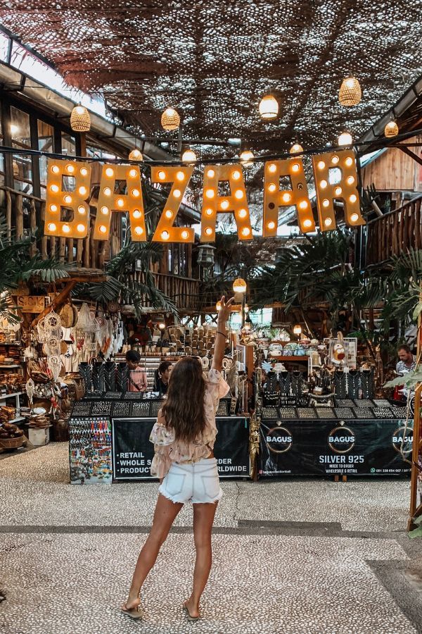 A woman in denim shorts and a floral top with her arm up in the air and a 'BAZAAR' sign hanging above a market in Bali, filled with artisanal goods and tropical decor.