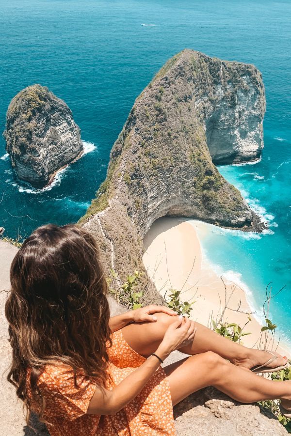 Overlooking the iconic Kelingking Beach in Nusa Penida, a woman in an orange dress sits on a high cliff edge, gazing at the turquoise waters and unique T-rex shaped coastline below.