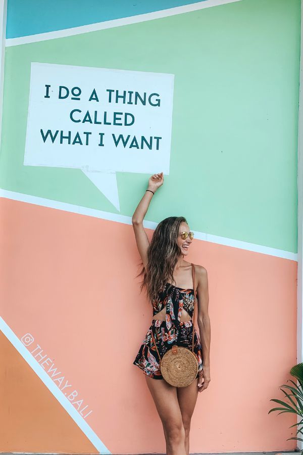 A woman in a floral set and round sunglasses raising her hand in front of a colorful wall with a speech bubble painting at The W Bali.