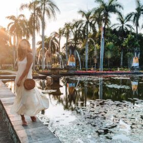 A woman in a white dress stands beside a tranquil pond with floating lotus flowers, backed by a serene row of palm trees and a glowing sunset sky.