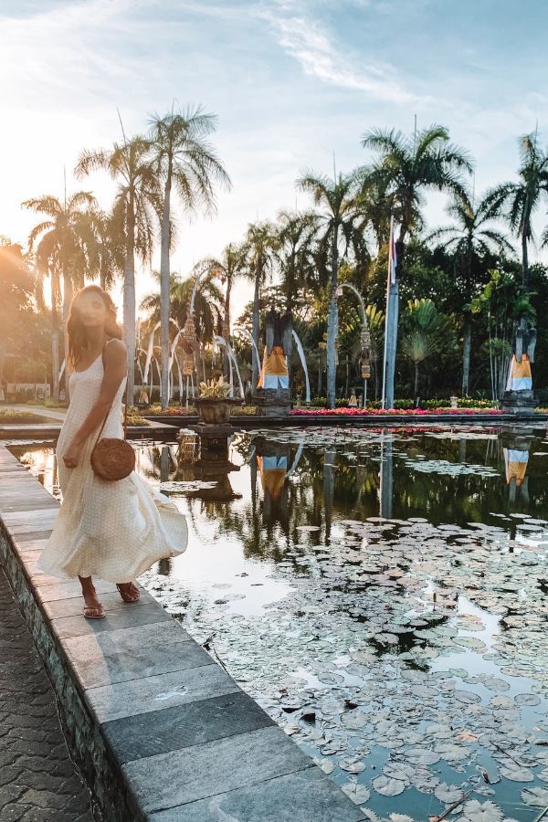 A woman in a white dress stands beside a tranquil pond with floating lotus flowers, backed by a serene row of palm trees and a glowing sunset sky.