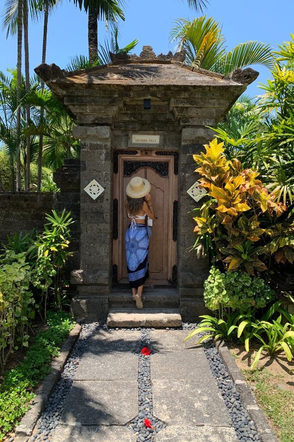A woman in a blue sarong and straw hat stands at the ornate doorway of a traditional Balinese villa, surrounded by lush tropical greenery and stone path with red hibiscus flowers.