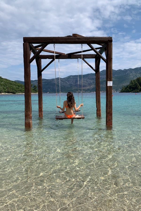 A woman in a swimsuit sitting on a wooden swing in crystal clear shallow water on an island on Croatia's Dalmatian coast.