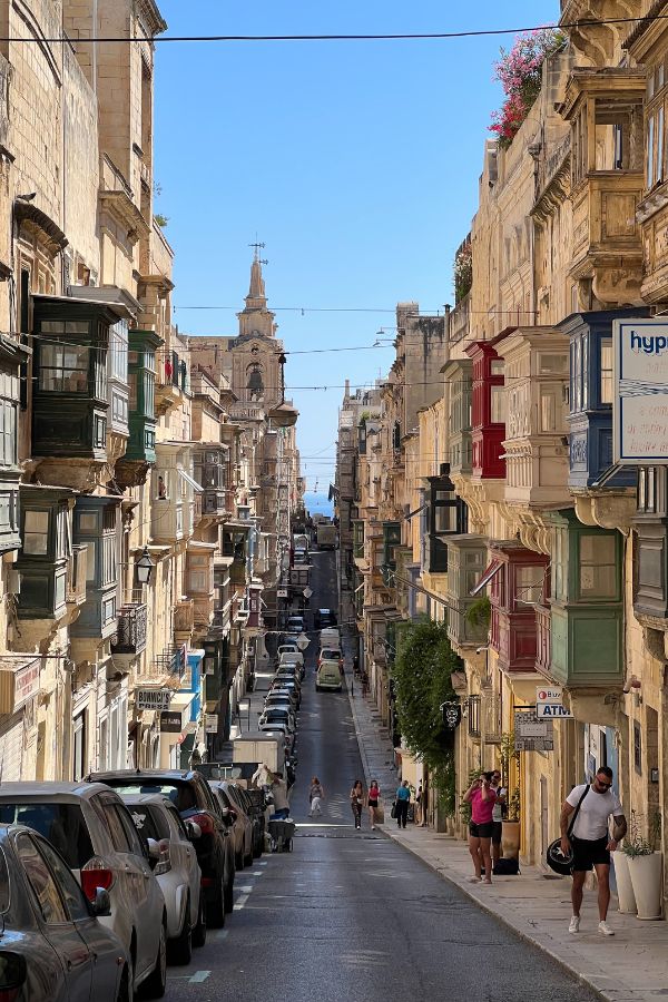 A hilly street in the heart of Valletta, Malta with stone buildings and colorful balconies on either side.