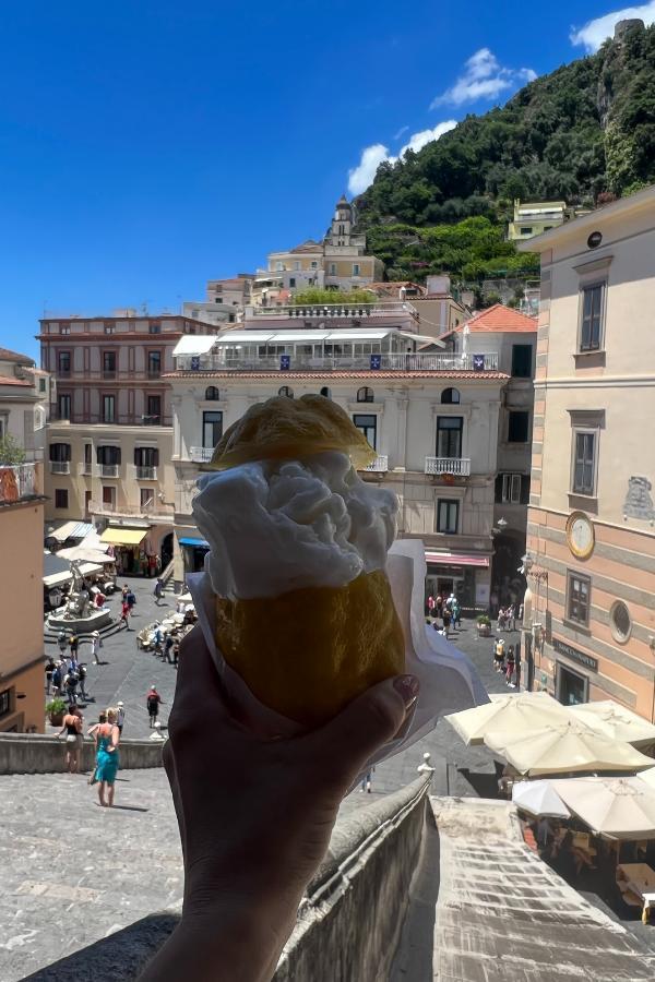 A hand holding a large lemon filled with traditional lemon sorbet from Amalfi in front of a bustling square along the Amalfi Coast.