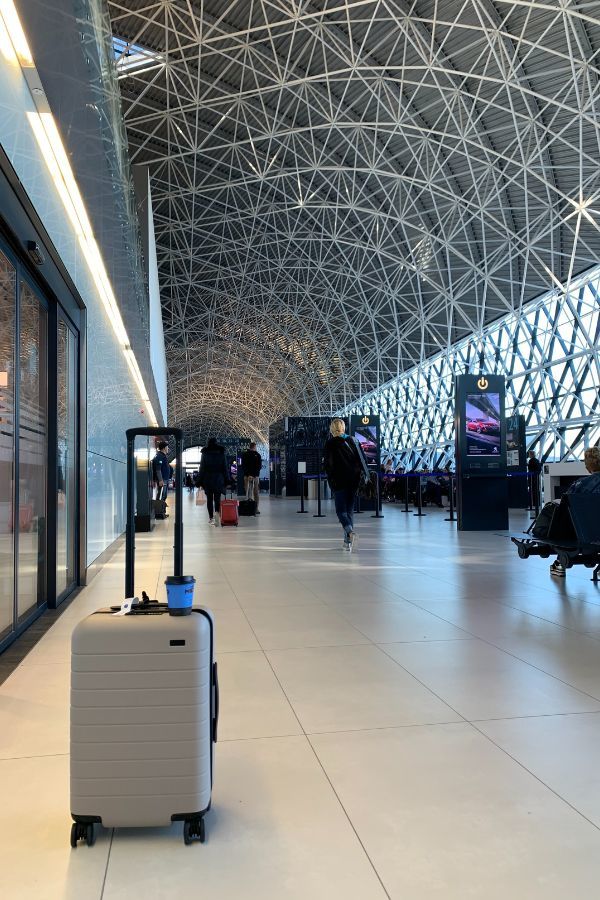 Beige carry on luggage in the middle of an airport with a blue to-go coffee cup sitting on top of it.